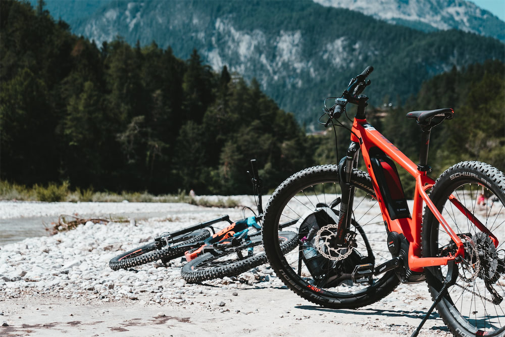 Due biciclette elettriche da mountain-bike di colore rosso e azzurro sulla riva di un torrente di montagna. Sullo sfondo panoramica sui versanti montuosi coperti di boschi di conifere.
