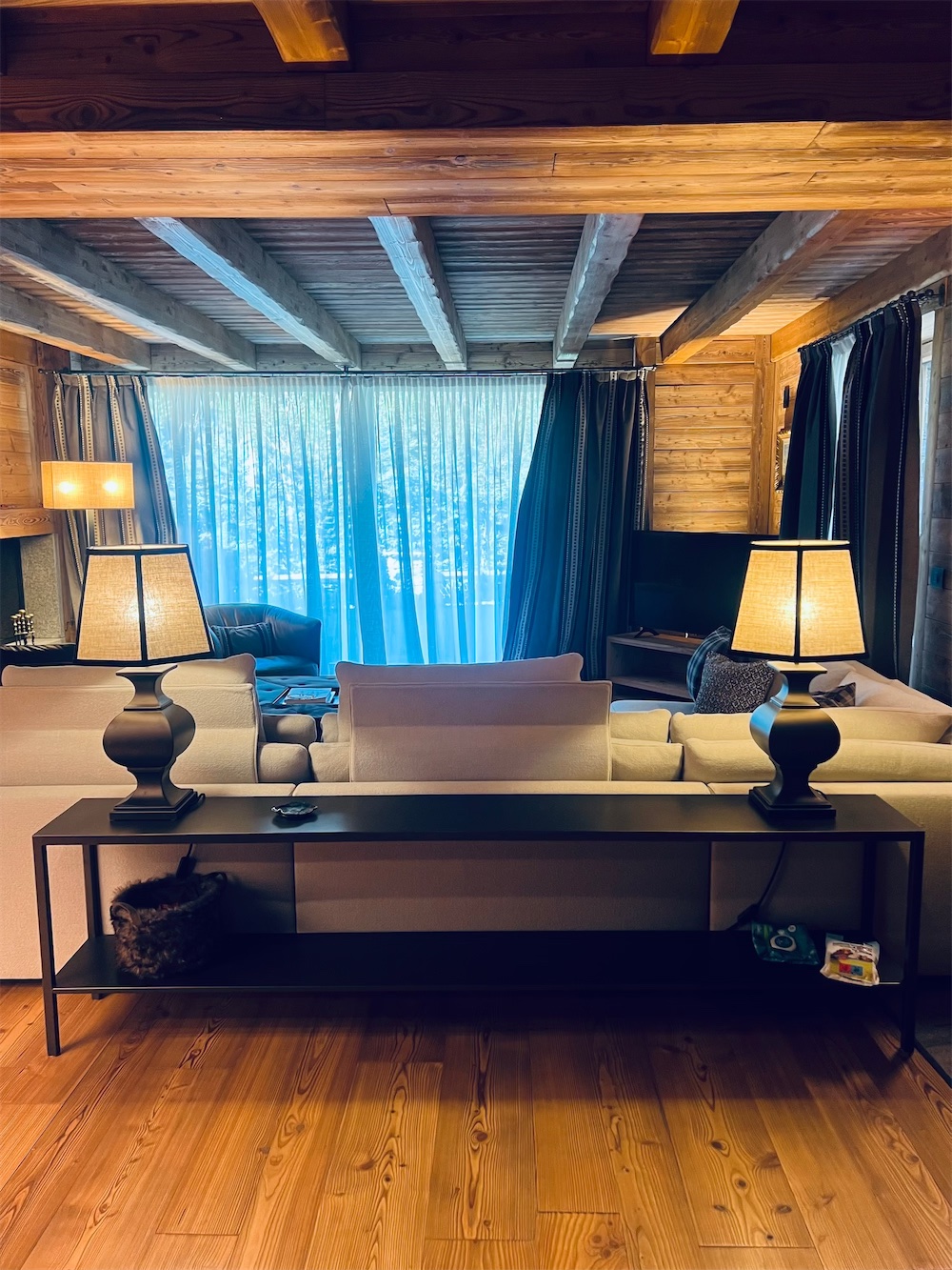 Case di lusso Aosta: scorcio di un soggiorno con parquet chiaro e soffitto in legno con travatura a vista. In primo piano un basso mobile a giorno marrone a due piani con una coppia di lampade, a base sagomata e paralume in pergamena chiaro, accese. Posteriormente, un ampio divano bianco prospiciente una portafinestra con tendoni azzurri.