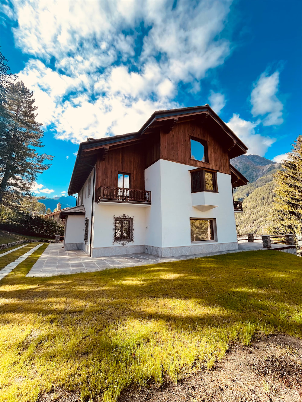 Case di lusso Aosta: vista di un edificio a due piani rivestito di legno e intonaco bianco, con quattro finestre rettangolari. Dietro la casa, montagne e cielo nuvoloso, in primo piano un prato verde su cui si stagliano ombre di nuvole.