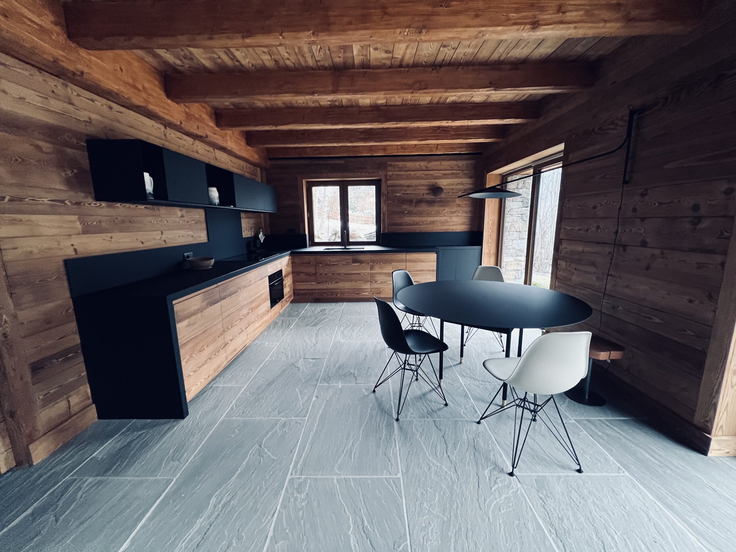 Immobili di prestigio Valle d'Aosta: interno di una cucina con pavimento di lastre di pietra grigia e pareti e soffitto in listoni di legno chiaro rustico. In primo piano un tavolo ovale nero con due sedie in plastica bianche e due nere con le gambe in metallo. Le basi sono in materiale nero con frontali in legno chiaro, i pensili completamente neri. Gli elettrodomestici sono incassati e il lavello si trova davanti a una finestra. Sulla destra in fondo una portafinestra.