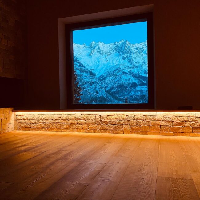 Particolare di una stanza con il pavimento in listoni di legno e un basso muretto in pietra lungo le pareti. Si apprezza il riquadro di una finestra a vetro unico che incornicia un paesaggio montuoso coperto di neve.