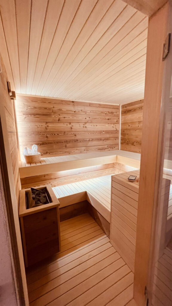 Locale sauna rivestito internamente di pannelli di legno naturale chiaro, completo di panche e braciere.