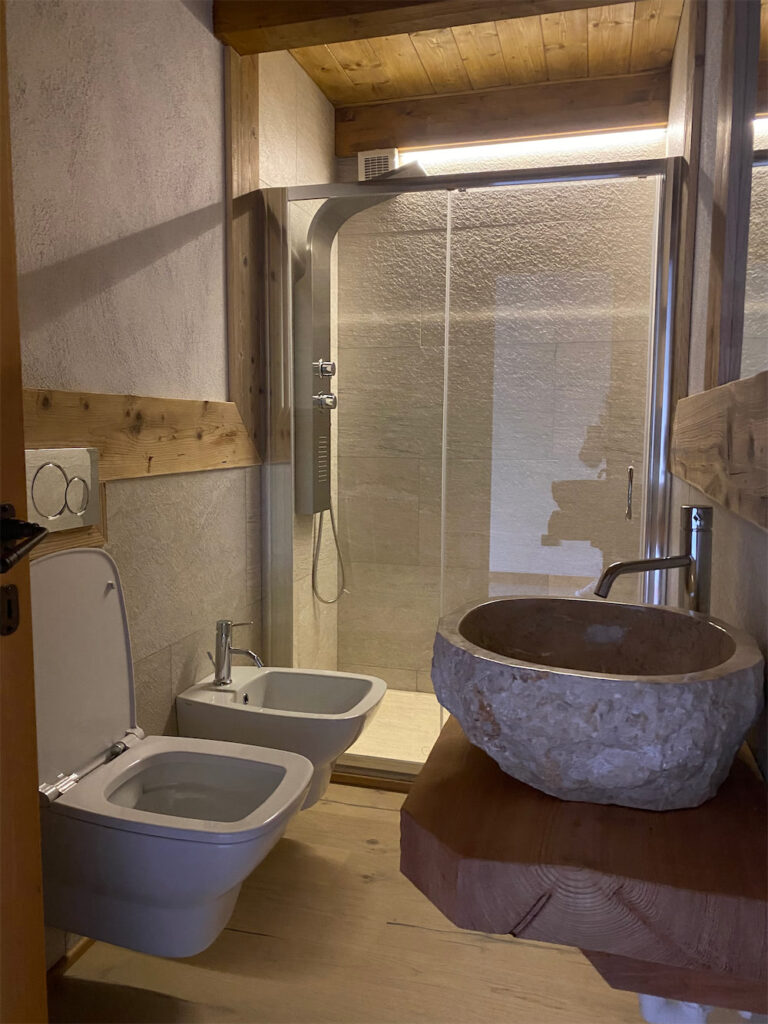 Stanza da bagno con vaso sanitario e bidet in ceramica bianca sospesi. In primo piano un lavabo in pietra grezza con rubinetto in acciaio, sullo sfondo piatto doccia chiuso da parete in vetro.