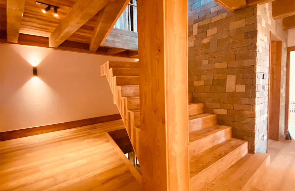 Locale mansarda con tetto a vista in legno biondo. Colonna portante centrale a sinistra di una scala in legno che porta a un abbaino, affiancata a destra da un muro in pietra.