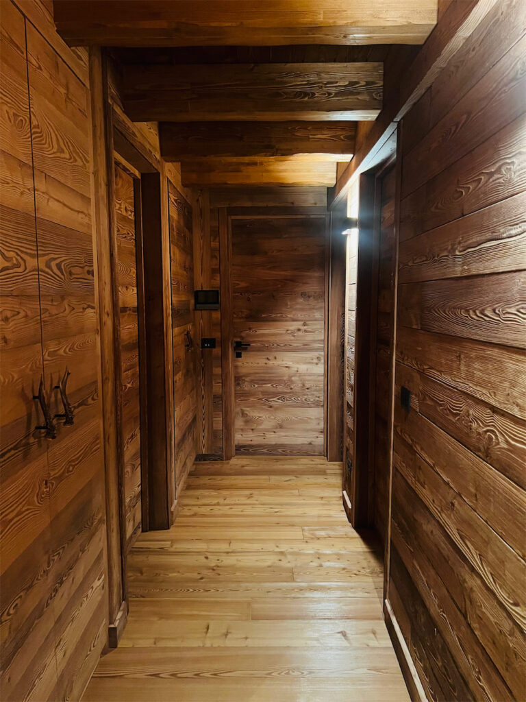Corridoio cieco completamente rivestito di legno con travi a vista. Porte in legno su entrambi i lati e sul fondo, a sinistra si notano le ante di un armadio a muro con maniglie fisse in metallo.