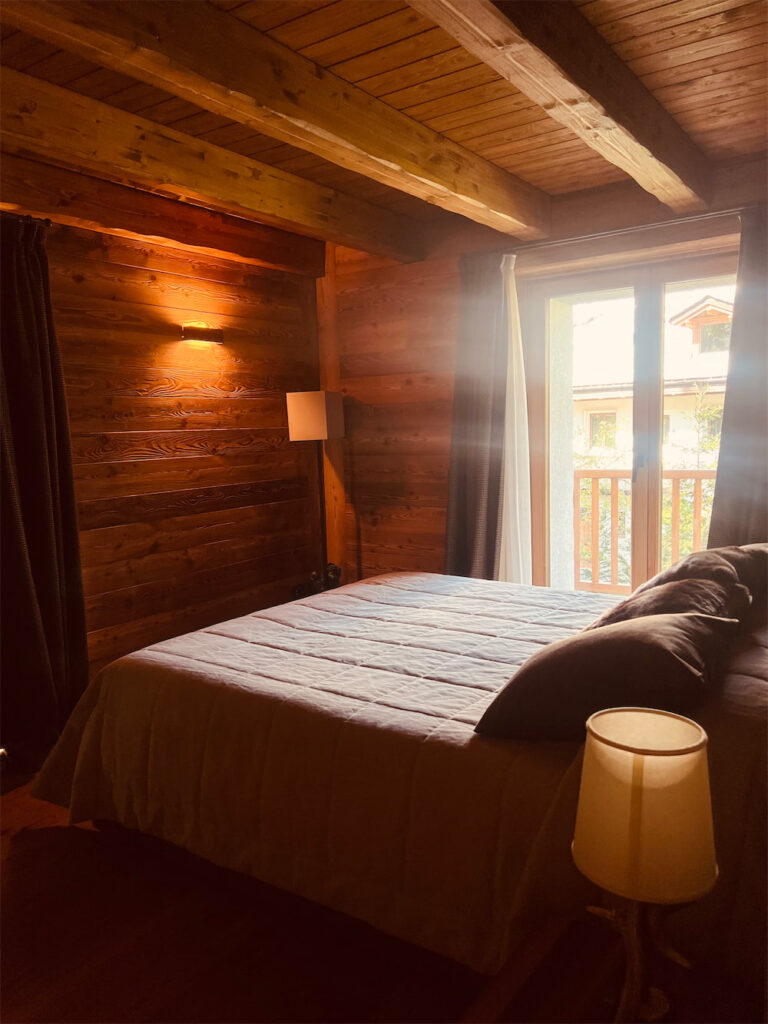 Camera da letto matrimoniale completamente rivestita in legno. In primo piano un letto a due piazze con copriletto beige chiaro e cuscini marroni, a fianco lampada da tavolo in tinta. Soffitto con travi a vista. Illuminazione a parete con doppia luce. Ampia portafinestra con accesso al balcone con ringhiera in legno.